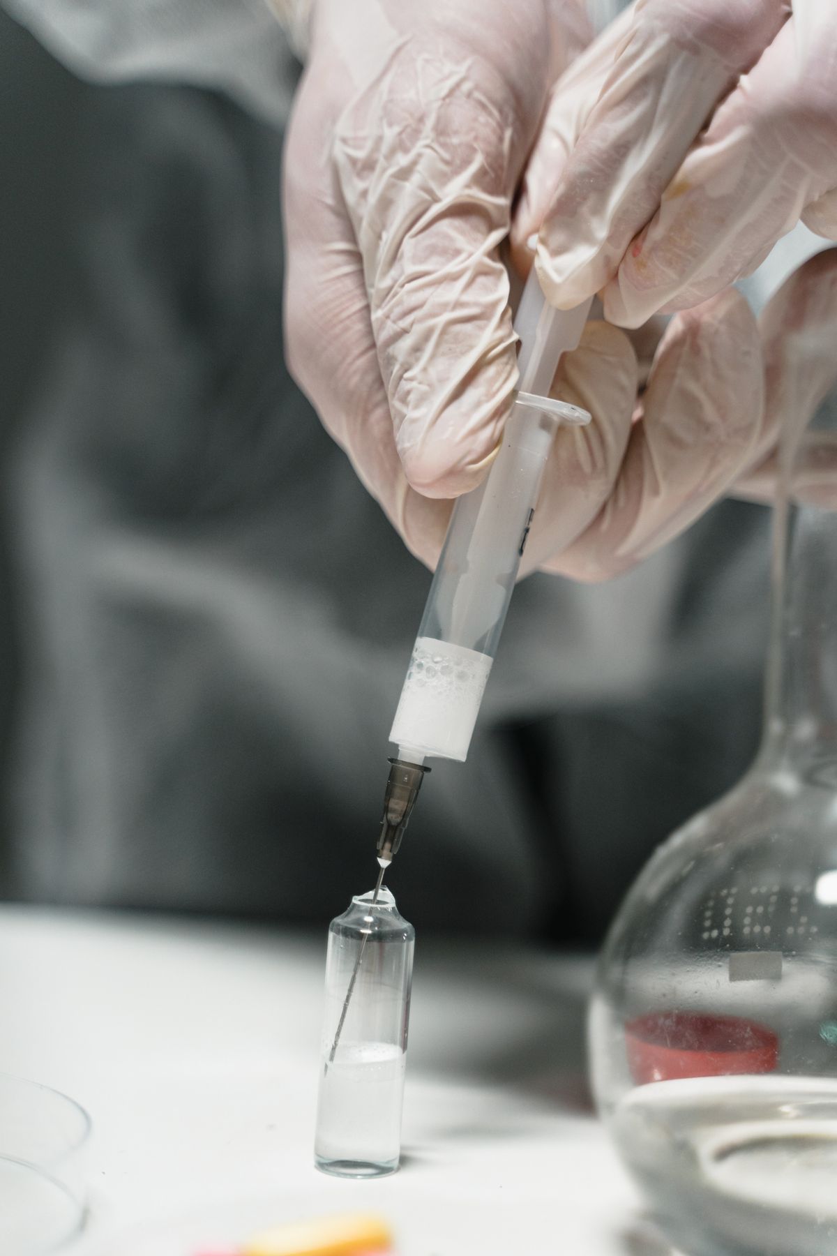 Gloved hands drawing liquid from a small glass vial with a syringe next to a laboratory flask