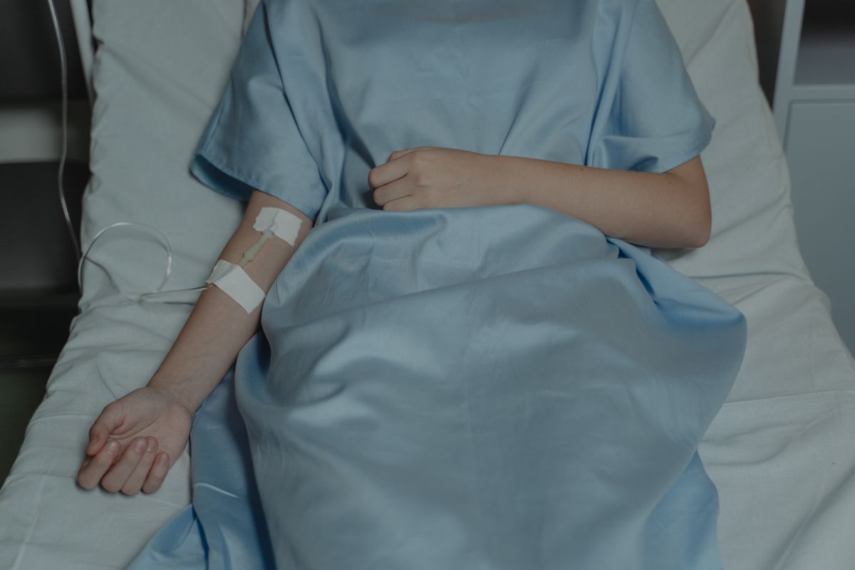A patient's forearm resting on a hospital bed with a cannula taped in place and tubing alongside